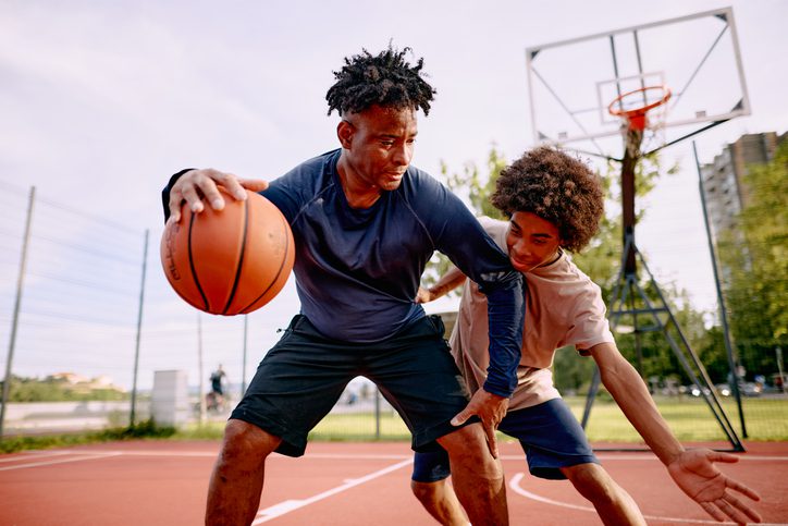 Father and son enjoying a sunny day while playing basketball on an outdoor court, sharing laughter and creating memories through active, healthy recreation together